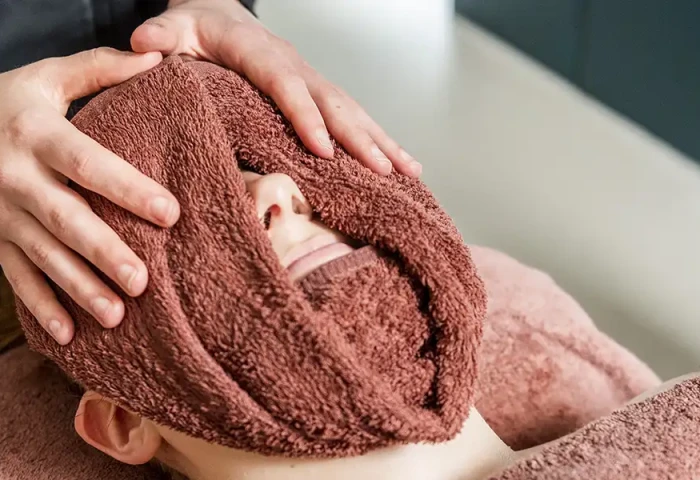 Client ligt in de stoel bij de schoonheidsspecialiste en heeft een warme handdoek om haar gezicht