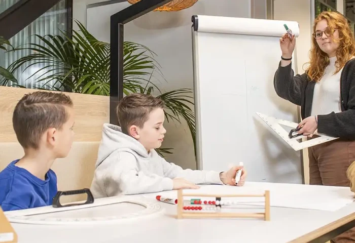 Een leerkracht staat voor kinderen en ze schrijft iets op een flipover. Er liggen telramen op tafel.
